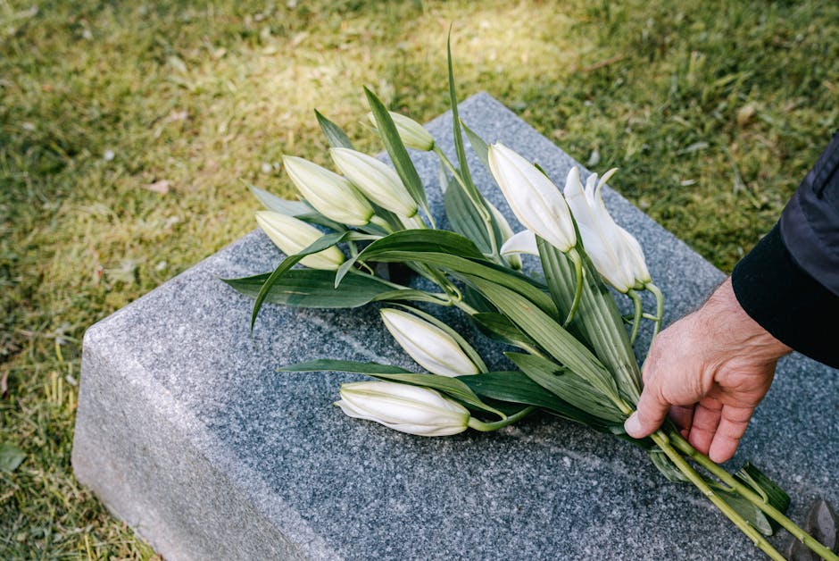 A person dressed in a dark coat is placing a bouquet of white lilies with long green leaves onto a grey granite headstone, set against a background of well-maintained green grass. The bouquet features
