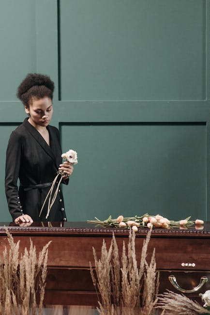 A woman with natural curly hair, dressed in a black blazer, stands solemnly next to a wooden casket adorned with a floral arrangement of pastel pink roses and white flowers. She holds a small white fl