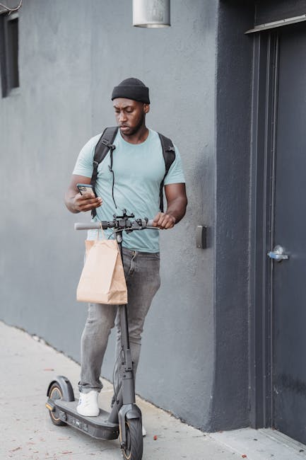 A young man dressed in a light blue t-shirt, grey jeans, and a black beanie is standing on an electric scooter while looking at his smartphone. He has a black backpack on and is holding a brown paper