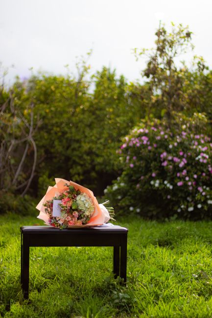 A vibrant bouquet of fresh flowers rests on a black wooden stool outdoors in a garden setting. The arrangement features pink roses, white hydrangeas, and small sprigs of greenery, wrapped in peach-col