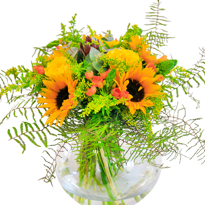 Expertly crafted by local Gidea Park florists, this bouquet is a visual melody of sunshine and community spirit. Towering sunflowers with luminescent yellow petals form the arrangement's centrepiece, their dark, inviting cores exuding warmth and happiness. Nestled among them are wispy yellow filler flowers, each adding depth and vibrancy that reflect the lush lawns and gardens of Gidea Park. Coral-orange cup blooms provide vivid splashes of colour, weaving through the greens for a subtle modern edge. The foliage is equally captivating-graceful ferns fan outward, soft eucalyptus sprigs introduce notes of silver-blue, and clusters of bright lime leaves infuse energy. Each stem, visible through the crystal-clear glass vase, suggests freshness and meticulous care, as if just gathered from local fields. The whole composition feels like a celebration of summer's arrival, radiating positivity and elegance. Whether presented as a centrepiece at a family gathering on Heath Drive or sent for same-day delivery, this bouquet embodies the joyful, neighbourly heart of Gidea Park.